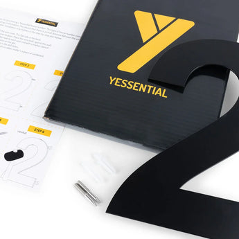 L'Adamo Black 30 cm de yessential.fr - un numéro 2 en métal noir frappant - comprend une feuille d'instructions, du matériel d'installation et une boîte noire avec le logo jaune Yessential, le tout soigneusement présenté sur une surface blanche.