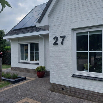 Une maison en briques blanches avec de grandes fenêtres, des panneaux solaires sur le toit, un numéro de maison Angelo Black de 30 cm yessential.fr (numéro 27) sur le mur, une plante en pot rouge à l'entrée et un trottoir carrelé à l'avant.