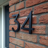 Installés sur un mur de briques rouges près d'une porte, les numéros de maison Milano Black 15 cm de yessential.fr créent un look moderne car leur design tridimensionnel projette des ombres audacieuses sur les briques.