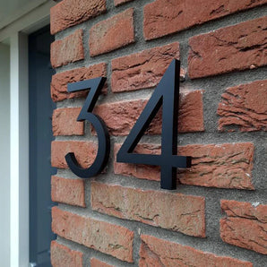 Installés sur un mur de briques rouges près d'une porte, les numéros de maison Milano Black 15 cm de yessential.fr créent un look moderne car leur design tridimensionnel projette des ombres audacieuses sur les briques.