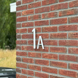 Les numéros de maison Amato White 20 cm de yessential.fr sont montés sur un mur de briques rouges, dans le style des numéros de maison modernes. Un peu de verdure et une partie d’une voiture noire apparaissent faiblement en arrière-plan.