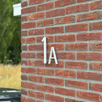 Les numéros de maison Amato White 20 cm de yessential.fr sont montés sur un mur de briques rouges, dans le style des numéros de maison modernes. Un peu de verdure et une partie d’une voiture noire apparaissent faiblement en arrière-plan.