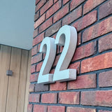 L'élégant numéro de maison Enrico béton 20 cm de yessential.fr est durable et monté sur un mur de briques rouges à côté d'une porte avec des panneaux en bois et une lampe carrée.