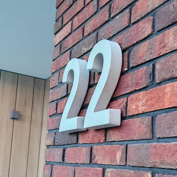 L'élégant numéro de maison Enrico béton 20 cm de yessential.fr est durable et monté sur un mur de briques rouges à côté d'une porte avec des panneaux en bois et une lampe carrée.