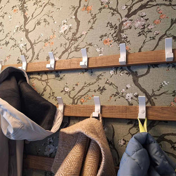 Deux porte-manteaux Huso White de yessential.fr sont fixés à un mur avec un motif floral. Des manteaux et une écharpe beige sont accrochés aux crochets, certains restent vides.