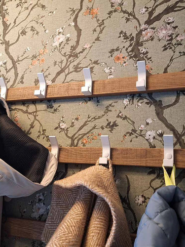 Deux porte-manteaux Huso White de yessential.fr sont fixés à un mur avec un motif floral. Des manteaux et une écharpe beige sont accrochés aux crochets, certains restent vides.