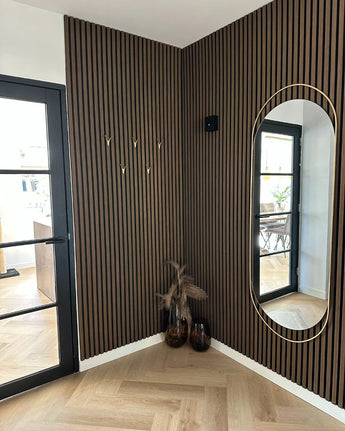 Une entrée moderne avec des murs verticaux à lattes de bois, un haut miroir ovale, un design minimaliste, 10x patères dorées Vilano Gold de yessential.fr, deux vases avec des plantes séchées, une porte vitrée et un sol à chevrons en bois clair.