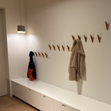 Un couloir minimaliste comprend des placards blancs, les crochets muraux en bois Fabio Oak de yessential.fr comme porte-manteau minimaliste en bois, un manteau beige, un sac sombre avec une section orange et une suspension qui éclaire l'espace.