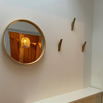 Un miroir rond avec un cadre en bois est accroché à un mur blanc et reflète une lampe cage à oiseaux et des rideaux orange. Trois patères en bois de yessential.fr Fabio Oak sont montées à côté du miroir au-dessus d'une armoire blanche.