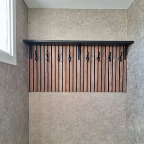 Le porte-manteau mural 10x Avano Black de yessential.fr comprend une étagère noire et des crochets élégants sur des lattes de bois verticales, présentés dans une pièce neutre avec une fenêtre à gauche.