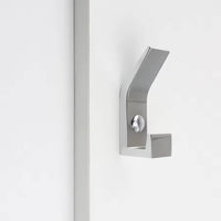 La patère Huso Chrome de yessential.fr présente une finition métallique brossée, un design minimaliste et des lignes angulaires nettes pour un look élégant, monté sur une surface blanche à côté d'une bande verticale.