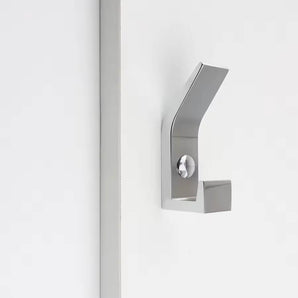 La patère Huso Chrome de yessential.fr présente une finition métallique brossée, un design minimaliste et des lignes angulaires nettes pour un look élégant, monté sur une surface blanche à côté d'une bande verticale.
