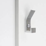 La patère Huso Chrome de yessential.fr présente une finition métallique brossée, un design minimaliste et des lignes angulaires nettes pour un look élégant, monté sur une surface blanche à côté d'une bande verticale.