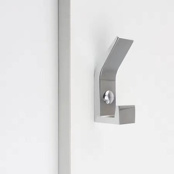La patère Huso Chrome de yessential.fr présente une finition métallique brossée, un design minimaliste et des lignes angulaires nettes pour un look élégant, monté sur une surface blanche à côté d'une bande verticale.