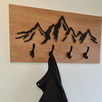 L'Avano Black de yessential.fr est un porte-manteau mural en bois avec quatre patères élégantes et un motif de chaîne de montagnes peint en noir. Un manteau sombre est suspendu à l'un des crochets.