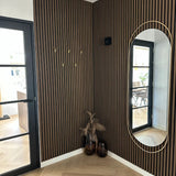 Une entrée moderne est dotée de murs verticaux à lattes de bois, d'un miroir ovale, de sols clairs à chevrons et d'élégants crochets Vilano Gold de yessential.fr. Des crochets dorés mettent en valeur deux vases au feuillage séché ; les portes en verre à cadre noir se reflètent dans le miroir.