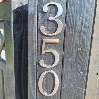 Les numéros de maison Matteo Nickel 10 cm de yessential.fr sont montés verticalement sur un poteau en bois sombre. Leur finition métallique brossée capte la lumière du soleil et projette des ombres qui mettent en valeur la texture du bois et l'éclat élégant des chiffres.