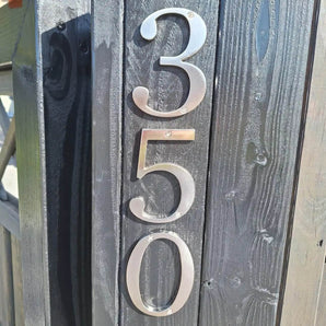 Les numéros de maison Matteo Nickel 10 cm de yessential.fr sont montés verticalement sur un poteau en bois sombre. Leur finition métallique brossée capte la lumière du soleil et projette des ombres qui mettent en valeur la texture du bois et l'éclat élégant des chiffres.
