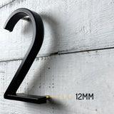 L'Amato Black 12,5 cm de yessential.fr est monté sur un mur en bois blanc avec un espace de 12 mm, ce qui montre clairement comment ce numéro de maison noir moderne se démarque de la surface.