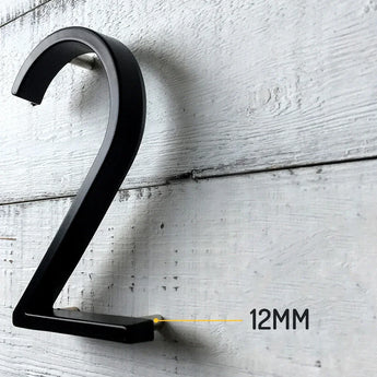 Le numéro de maison Amato Noir 15 cm de yessential.fr, numéro en métal 2, est monté en hauteur sur un mur en bois blanc. Une ligne jaune accentue l'espacement de 12 mm et souligne le design moderne.
