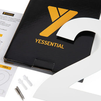 Un grand numéro de maison Adamo White 15 cm 2 de yessential.fr recouvre partiellement une boîte noire avec un logo jaune. Les fiches d'instructions, les chevilles et les vis se trouvent à proximité sur une surface blanche.