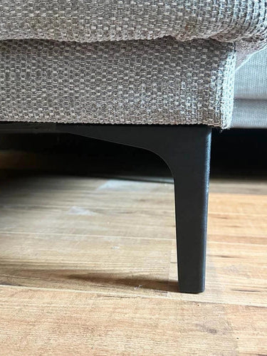 Gros plan d'un canapé texturé beige avec des pieds yessential.fr Menno Black de 15 cm sur un plancher en bois, mettant en valeur l'élégant pied en acier noir et les détails en tissu.