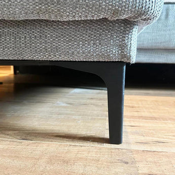 Gros plan d'un canapé texturé beige avec des pieds yessential.fr Menno Black de 15 cm sur un plancher en bois, mettant en valeur l'élégant pied en acier noir et les détails en tissu.