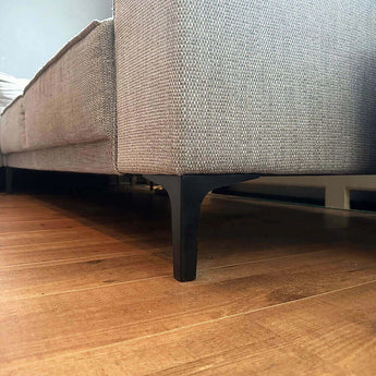 Gros plan d'un canapé en tissu gris avec des pieds de meuble en acier noir yessential.fr Menno Black de 13 cm sur un plancher en bois, en mettant l'accent sur la zone située sous le canapé.