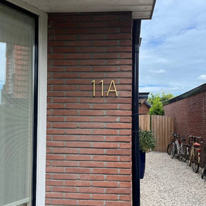 Un mur extérieur en brique est équipé de numéros de maison Amato Gold de 12,5 cm de yessential.fr. En arrière-plan, un chemin de gravier, une clôture en bois, des plantes en pot et des vélos garés apportent un charme supplémentaire.