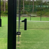 Une porte vitrée avec un grand numéro de maison Animo Black de 15 cm « 1 » de yessential.fr (sans perceuse) s'ouvre sur un court de paddle-tennis extérieur vert avec filet, entouré d'arbres et de clôtures grillagées.