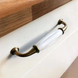 Un gros plan de la poignée Daro Ceramic Antique Gold de yessential.fr, avec de l'or antique et de la céramique blanche sur un tiroir au dessus en bois - idéale pour les fans de ferrures de meubles classiques.