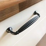 Un gros plan de la poignée de tiroir Alvaro Anthracite de yessential.fr, avec un design noir nervuré, montée sur une porte d'armoire blanche avec un comptoir en bois au-dessus.