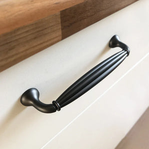 Un gros plan de la poignée de tiroir Alvaro Anthracite de yessential.fr, avec un design noir nervuré, montée sur une porte d'armoire blanche avec un comptoir en bois au-dessus.