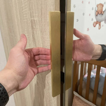 Les mains d'une personne s'étendent pour ouvrir une armoire équipée de poignées Enzo Gold de yessential.fr ; une crèche en bois et un mur avec des décorations d'étoiles et d'ours en peluche apparaissent en arrière-plan.