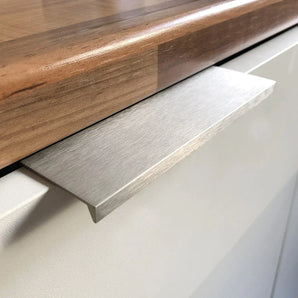 Gros plan d'une porte d'armoire blanche avec la poignée Enzo en acier inoxydable de yessential.fr, dotée d'une finition brossée élégante et d'un design rectangulaire plat, fini avec un comptoir en bois pour un look élégant et moderne.