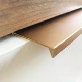Gros plan sur la bande décorative Enzo Rose Gold de yessential.fr, au profil en L et à la finition lisse brossée, installée en bordure d'un mur blanc sous du bois - idéale à associer avec des poignées de meubles modernes pour un intérieur stylé.