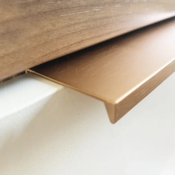 Gros plan sur la bande décorative Enzo Rose Gold de yessential.fr, au profil en L et à la finition lisse brossée, installée en bordure d'un mur blanc sous du bois - idéale à associer avec des poignées de meubles modernes pour un intérieur stylé.