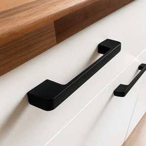 Gros plan des poignées Ramiro Black Deluxe de yessential.fr sur une armoire blanche avec un comptoir en bois. Ces poignées luxueuses rehaussent le design minimaliste et contemporain.