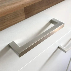 Gros plan d'une poignée carrée Vasco moderne en acier inoxydable de yessential.fr en acier inoxydable brossé sur une armoire blanche avec un dessus en bois au-dessus. Une autre poignée en acier inoxydable est visible en arrière-plan.