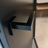 Un gros plan de la poignée de porte moderne Domaso Black de yessential.fr, avec une base carrée et un design angulaire minimaliste, montée sur une porte sombre avec la pièce légèrement décolorée visible derrière elle.