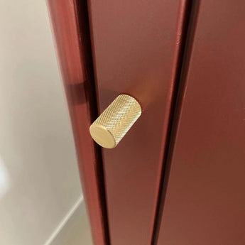 Un gros plan du bouton Remo Gold Deluxe de yessential.fr, un bouton de meuble de luxe en or avec un design cylindrique texturé, présenté sur une armoire ou un tiroir rouge foncé mat.