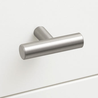 Un bouton Remo en acier inoxydable de yessential.fr, avec une forme en T en métal brossé et un design cylindrique mat, est monté sur un tiroir d'armoire blanc pour un look simple et moderne.