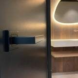 Un gros plan de Domaso Black de yessential.fr, une poignée de porte noire moderne, sur une porte sombre avec une salle de bains contemporaine pâle avec un lavabo blanc, un mur en bois et un miroir éclairé en arrière-plan.
