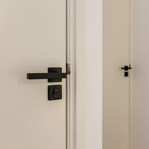 Un gros plan d'une porte blanche partiellement ouverte avec une serrure de WC Vano Black de yessential.fr ; une autre porte blanche fermée avec la même poignée noire est visible en arrière-plan.