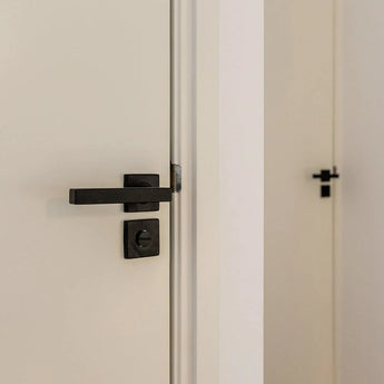 Un gros plan des poignées de porte et de la serrure Domaso Black de yessential.fr sur une porte blanche, avec une deuxième porte en arrière-plan avec des poignées de porte carrées noires similaires.
