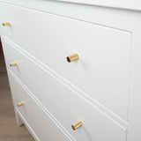 Gros plan d'une commode blanche avec trois tiroirs, chacun équipé d'un bouton Remo Gold Deluxe de yessential.fr. Les poignées cylindriques dorées ajoutent une touche d'élégance lorsque le buffet repose sur un parquet en bois clair.