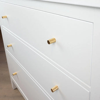 Gros plan d'une commode blanche avec trois tiroirs, chacun équipé d'un bouton Remo Gold Deluxe de yessential.fr. Les poignées cylindriques dorées ajoutent une touche d'élégance lorsque le buffet repose sur un parquet en bois clair.