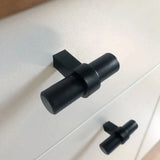Gros plan de deux boutons noirs Arturo de yessential.fr dans un style de cuisine moderne à boutons en T, montés sur une armoire blanche. La conception cylindrique comporte une barre horizontale sur une base verticale pour un look élégant et contemporain.