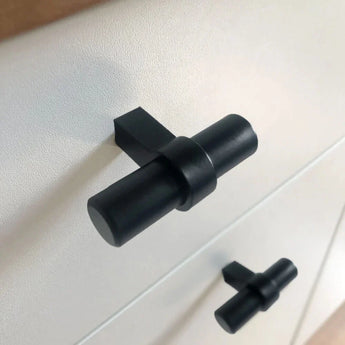 Gros plan de deux boutons noirs Arturo de yessential.fr dans un style de cuisine moderne à boutons en T, montés sur une armoire blanche. La conception cylindrique comporte une barre horizontale sur une base verticale pour un look élégant et contemporain.