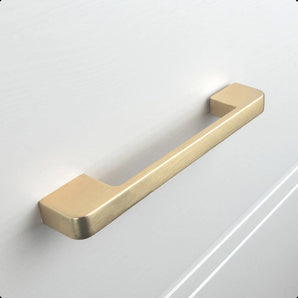 Une poignée d'armoire de cuisine Ramiro Gold Deluxe de yessential.fr, avec une finition or mat, est montée sur un tiroir d'armoire blanc avec une surface lisse et des rainures subtiles.